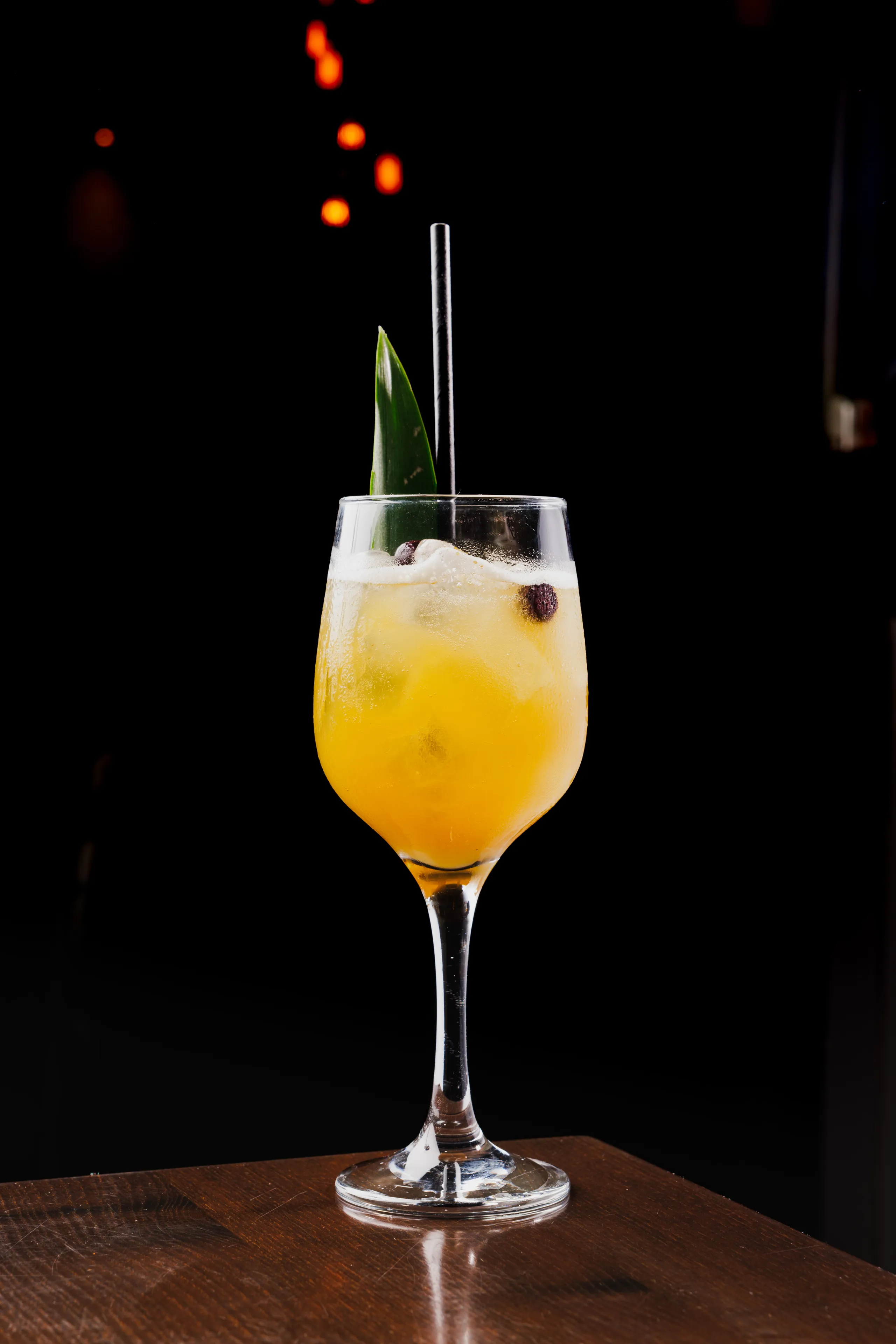 Yellow tropical cocktail in a stemmed glass with pineapple leaf garnish and berries on a dark bar background