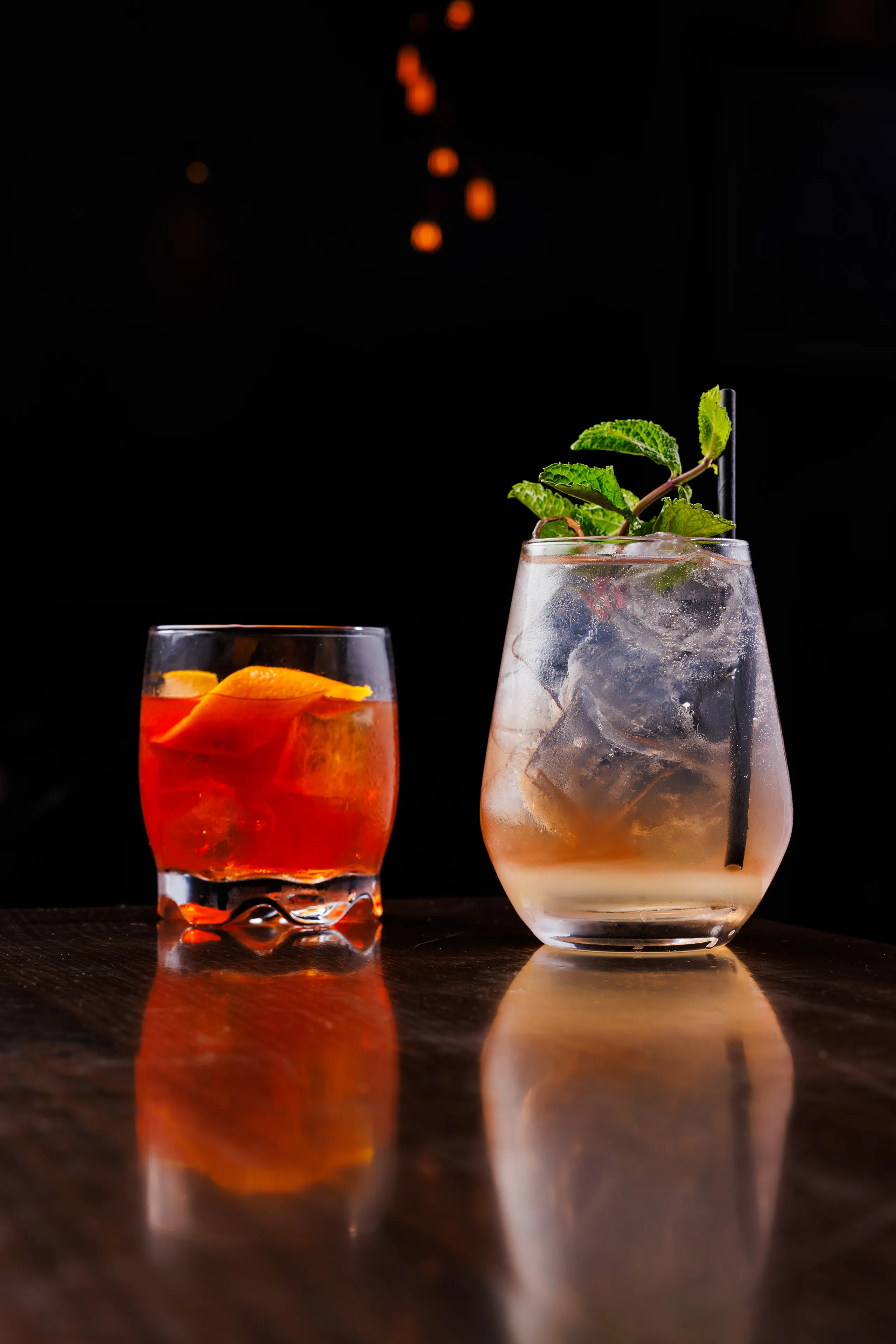 Two cocktails on a dark reflective bar, one garnished with orange peel and the other with mint leaves and ice.