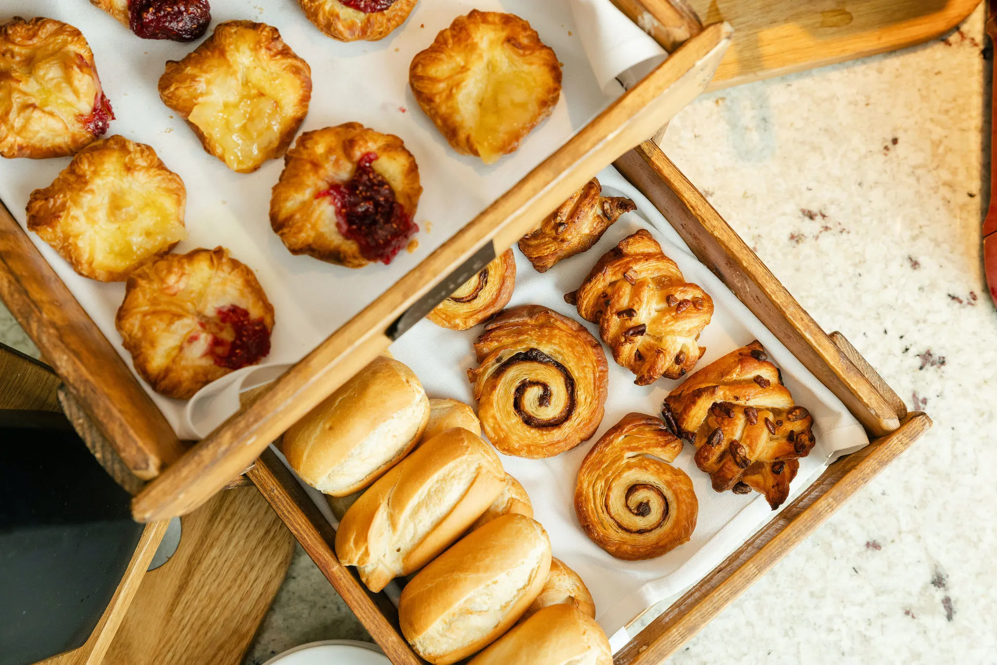 Assorted freshly baked pastries and bread rolls arranged in wooden trays on a buffet counter.