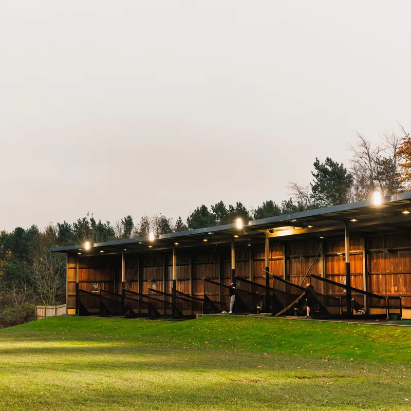 Illuminated golf driving range bays beside a green practice field at dusk