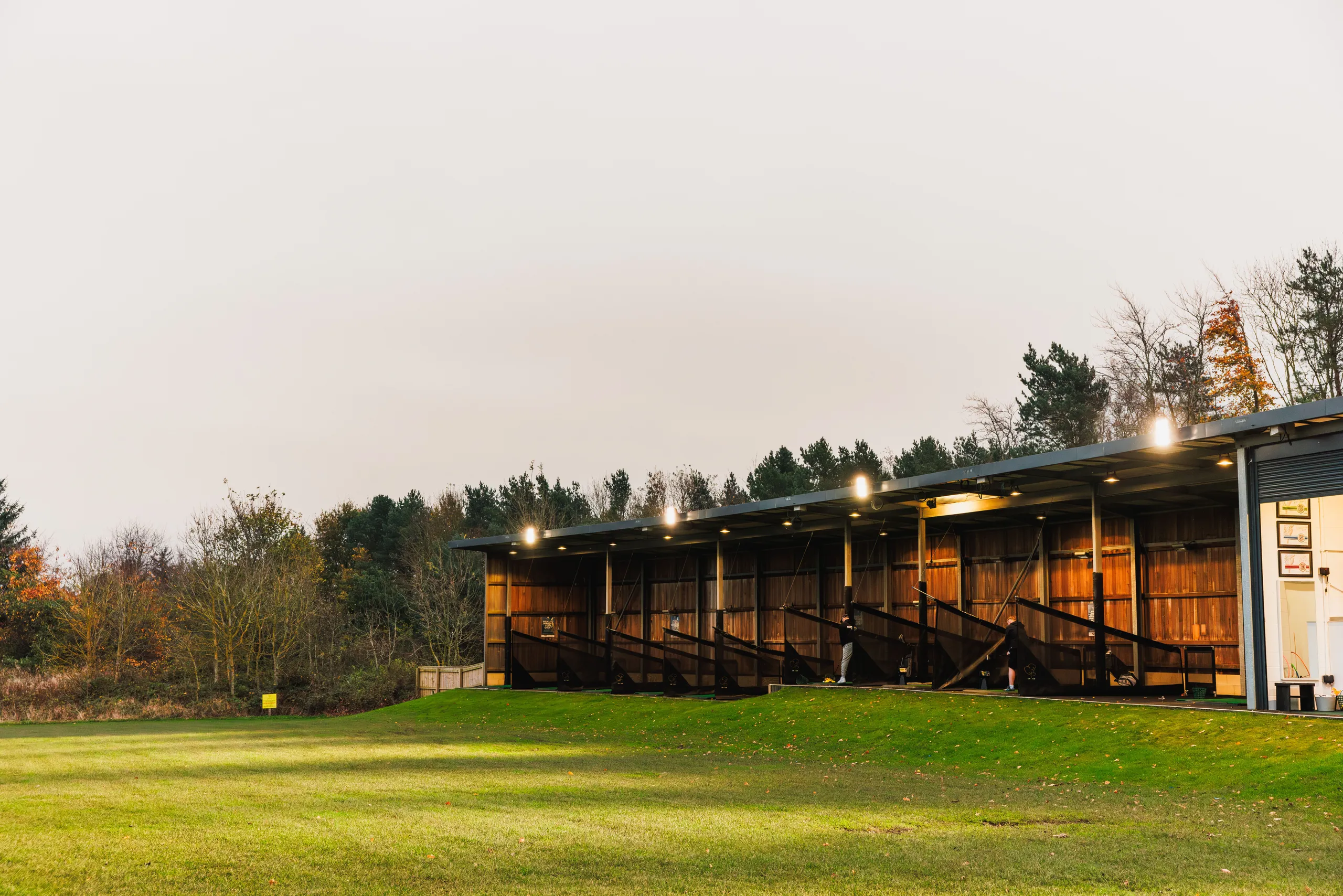 Illuminated golf driving range bays beside a green practice field at dusk