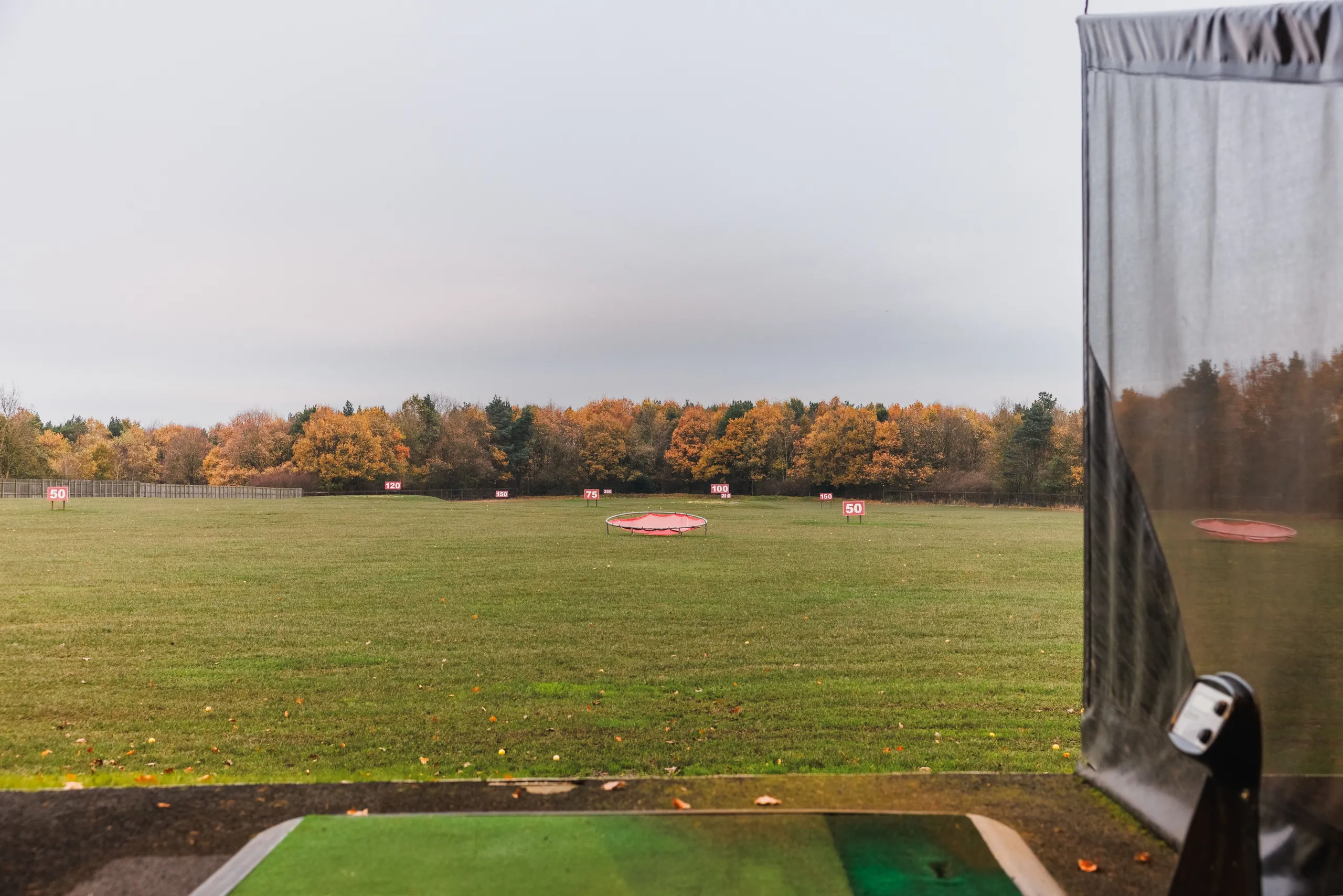 Outdoor golf driving range with distance markers, a target net and autumn trees under an overcast sky.