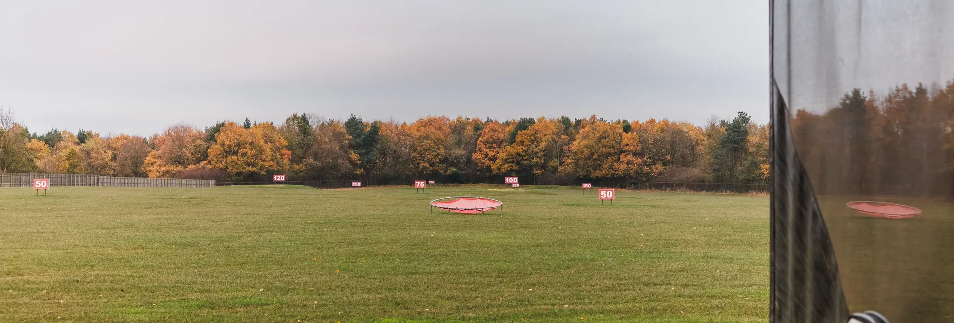 Outdoor golf driving range with distance markers, a target net and autumn trees under an overcast sky.