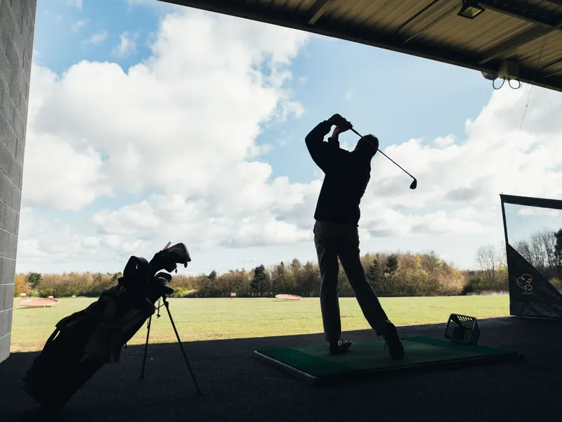 Silhouetted golfer finishing a swing at a covered driving range beneath a bright cloudy sky.