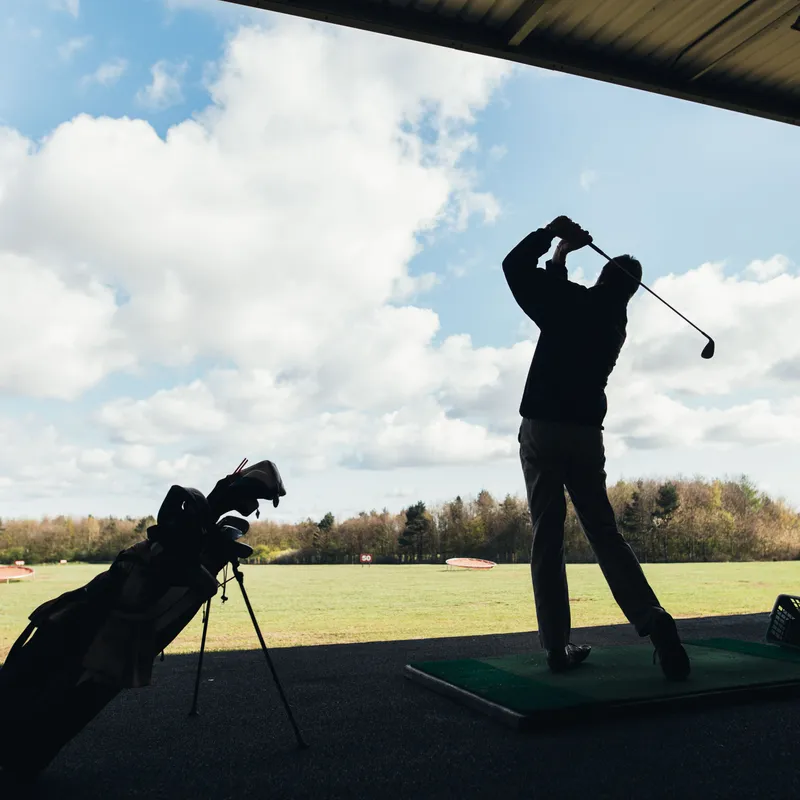 Silhouetted golfer finishing a swing at a covered driving range beneath a bright cloudy sky.