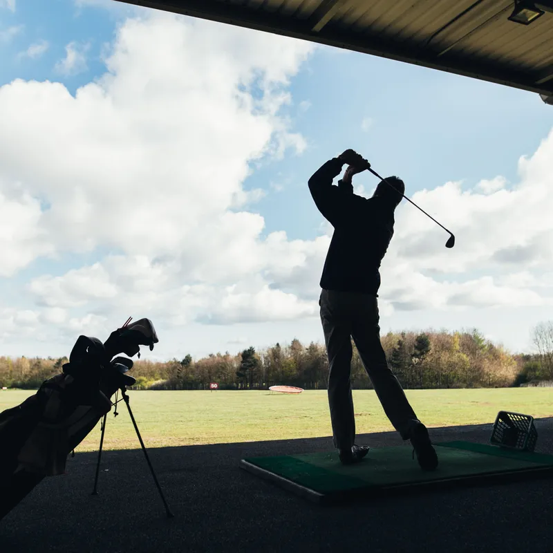 Silhouetted golfer finishing a swing at a covered driving range beneath a bright cloudy sky.