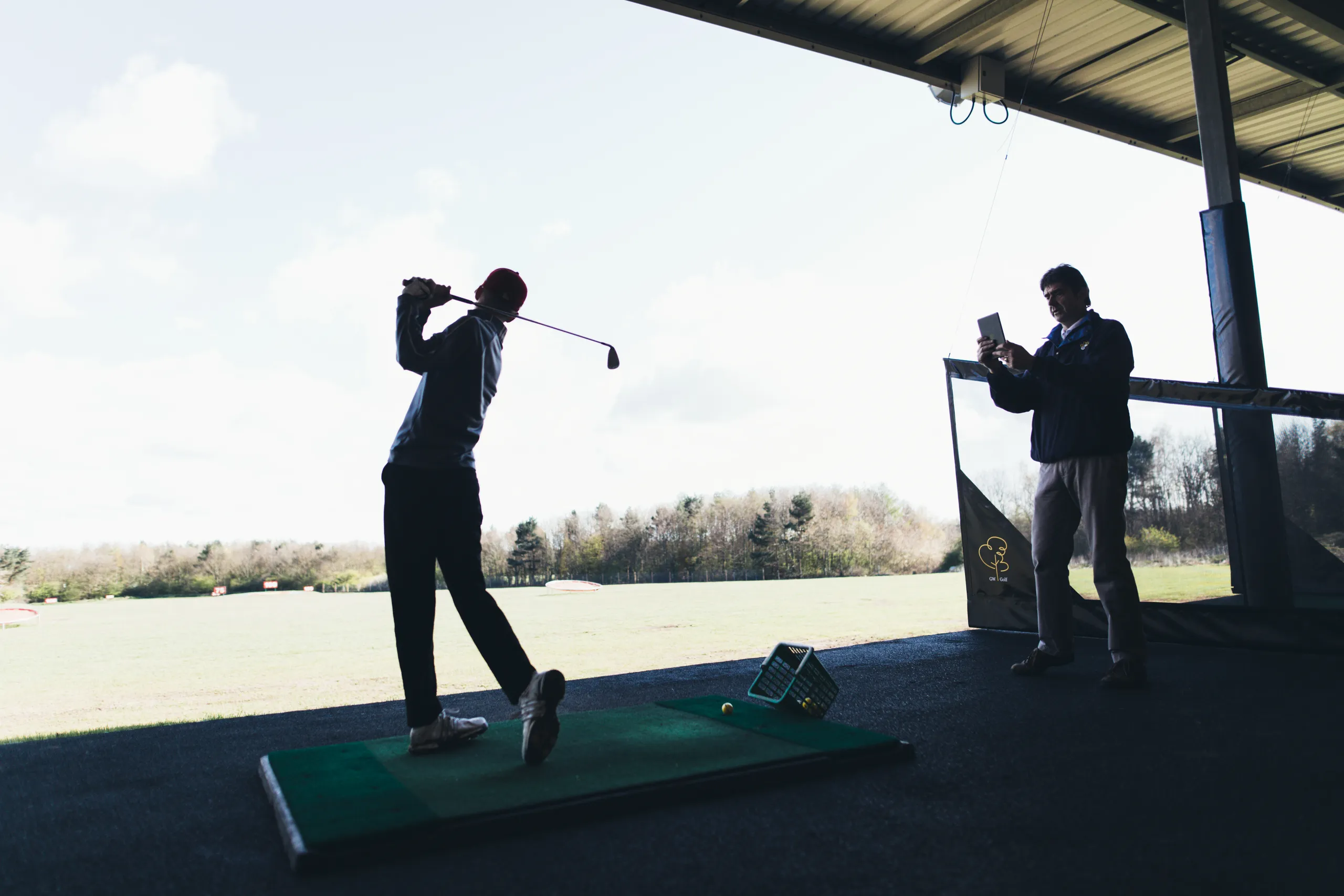 Golfer practising a swing at a covered driving range while another man records on a tablet.