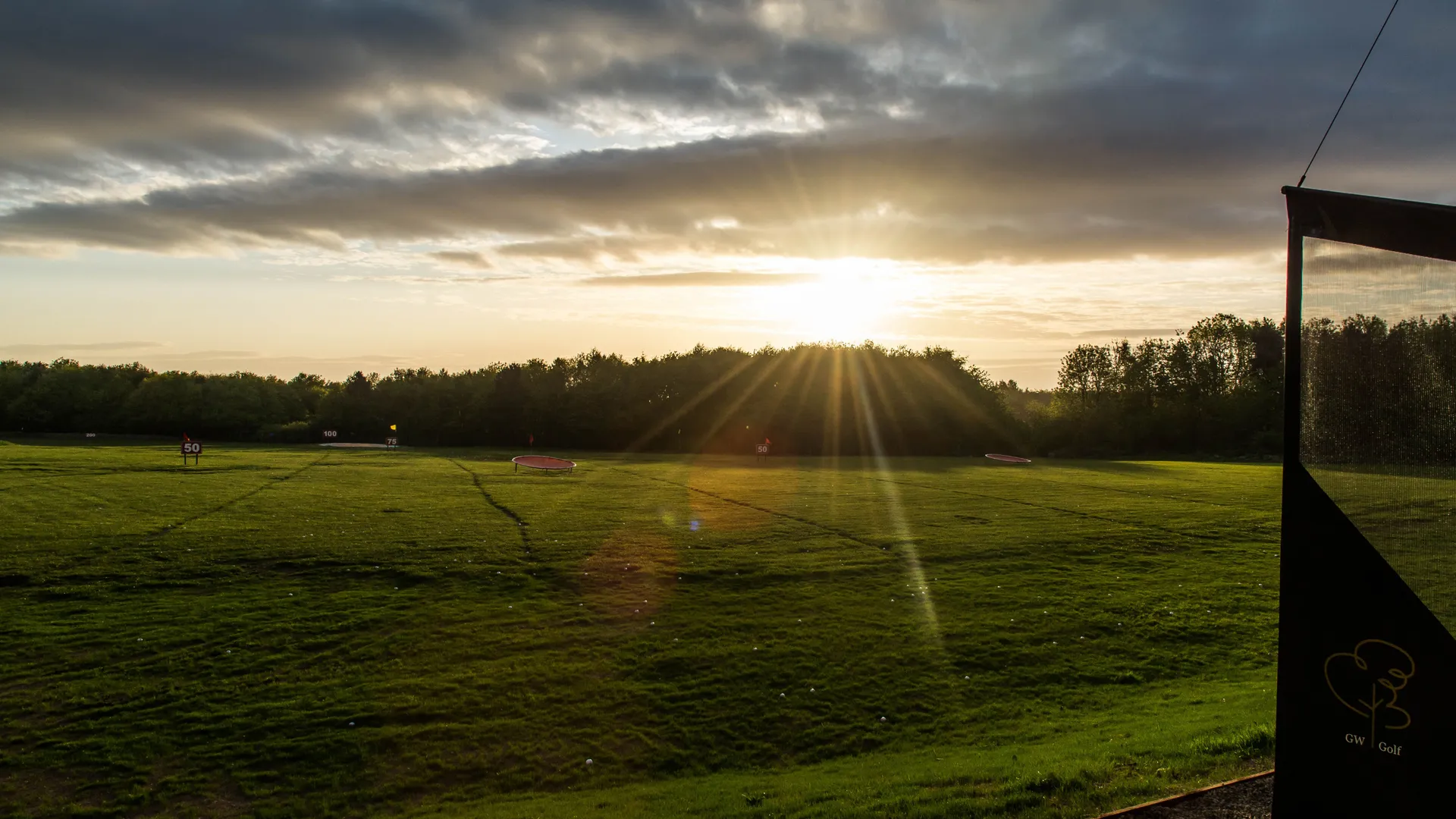 Sunrise over a golf driving range, with golden rays across the grass and dark clouds overhead.