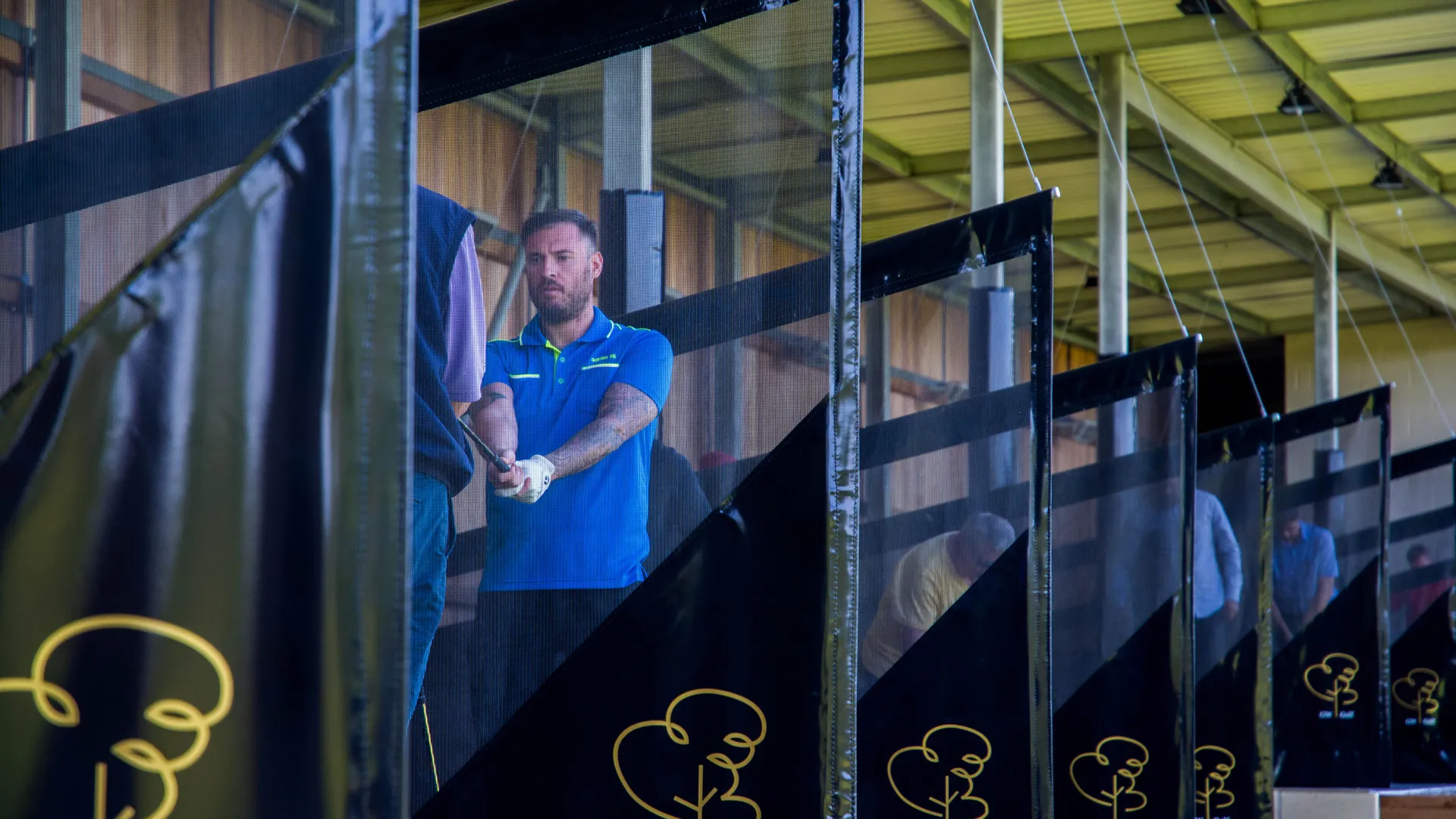 Man in a blue polo shirt practising his golf swing in a covered driving range bay
