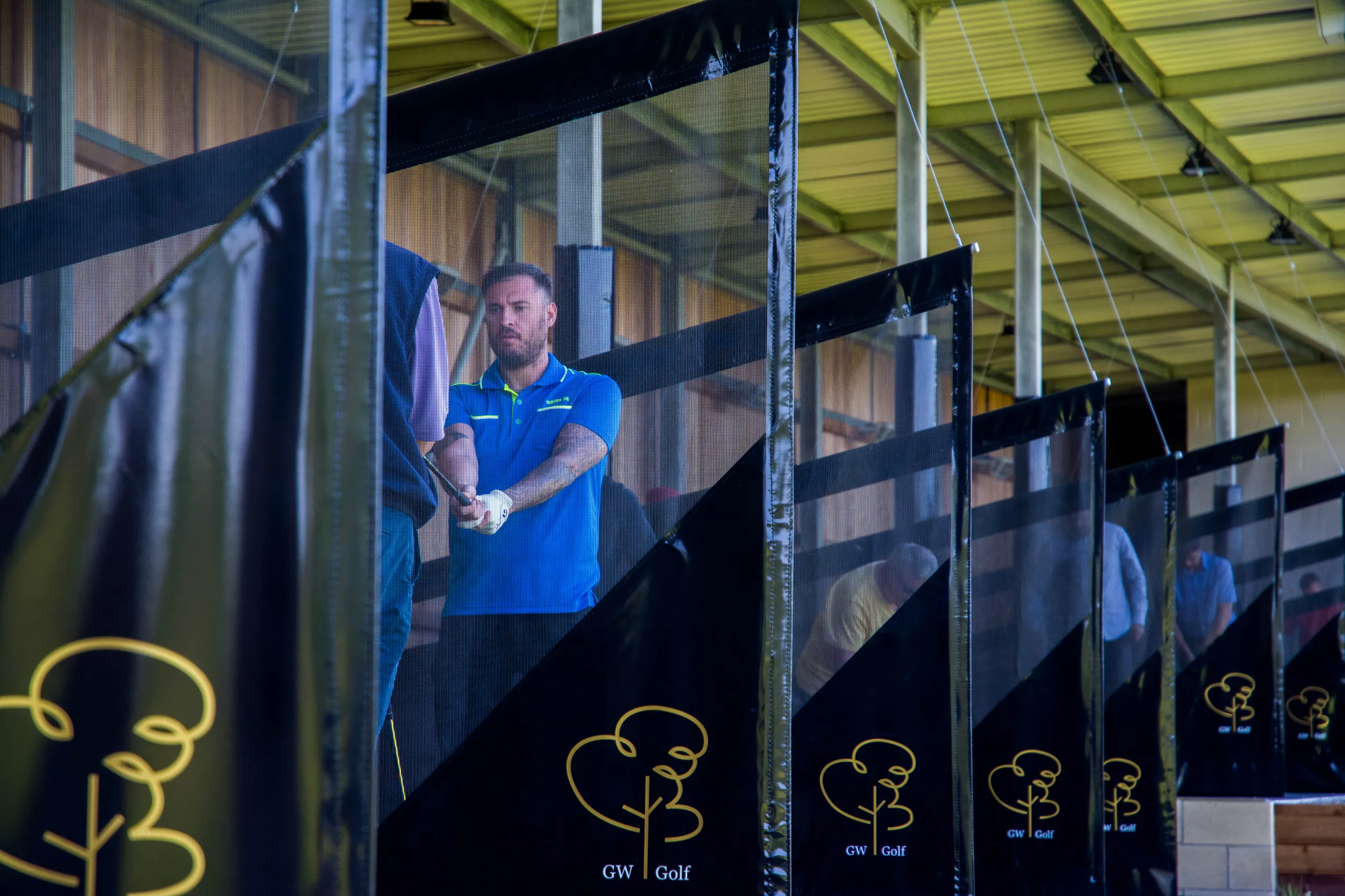 Man in a blue polo shirt practising his golf swing in a covered driving range bay