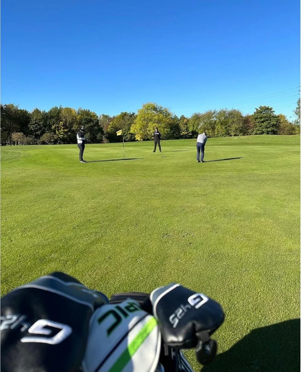 Three golfers on a green beside a flag, viewed over a golf bag under a clear blue sky.