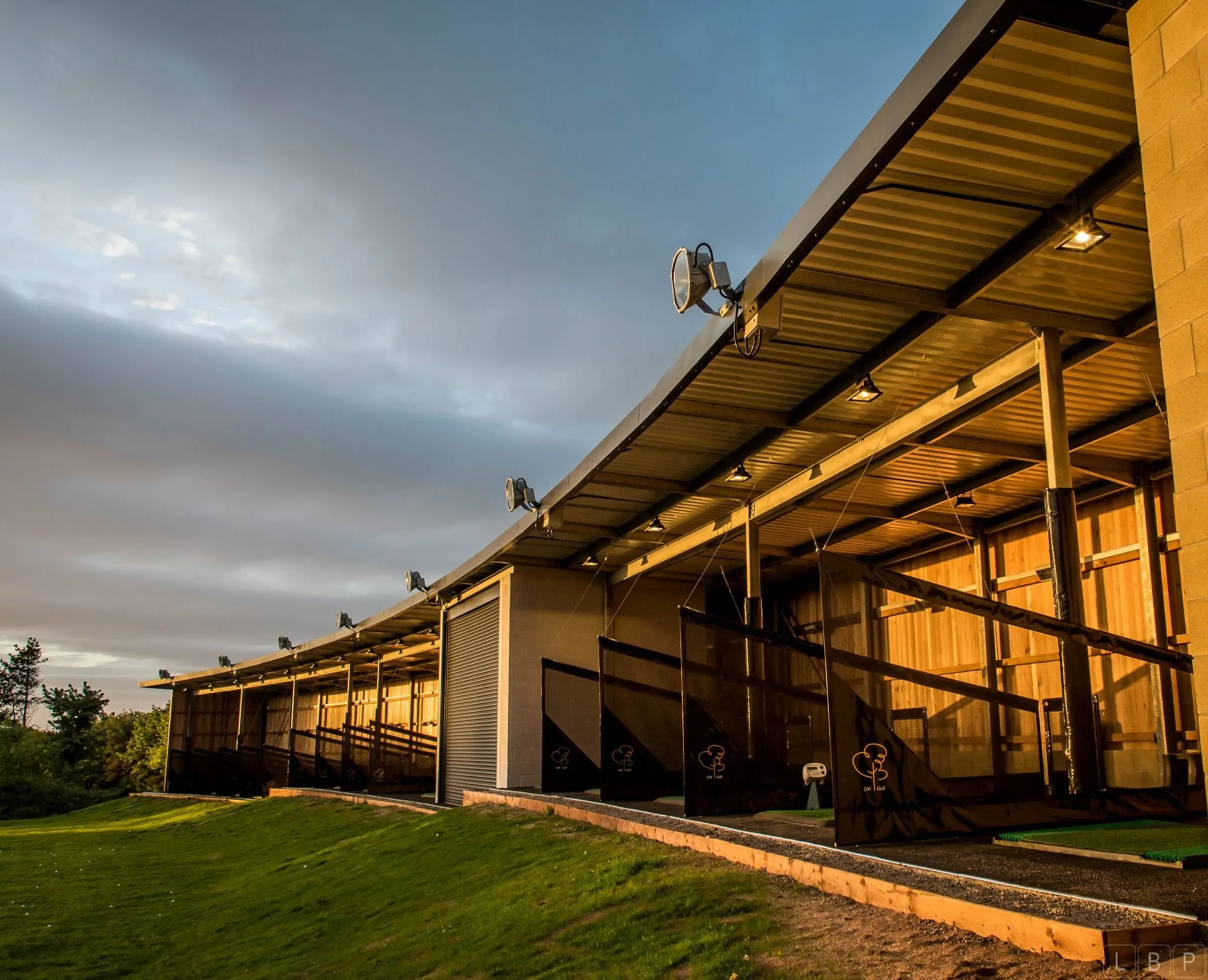 Covered golf driving range bays illuminated by warm evening light beneath a dramatic cloudy sky.