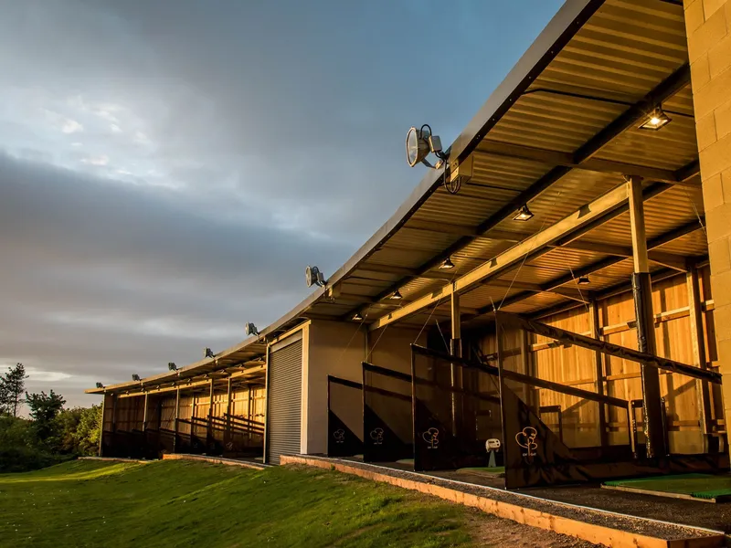 Covered golf driving range bays illuminated by warm evening light beneath a dramatic cloudy sky.