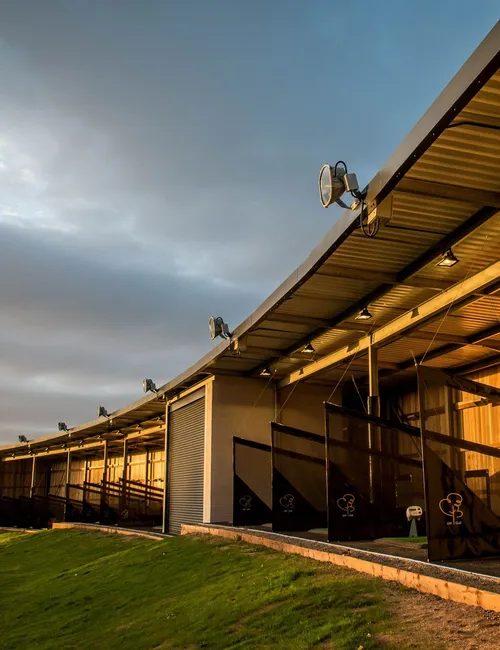 Covered golf driving range bays illuminated by warm evening light beneath a dramatic cloudy sky.