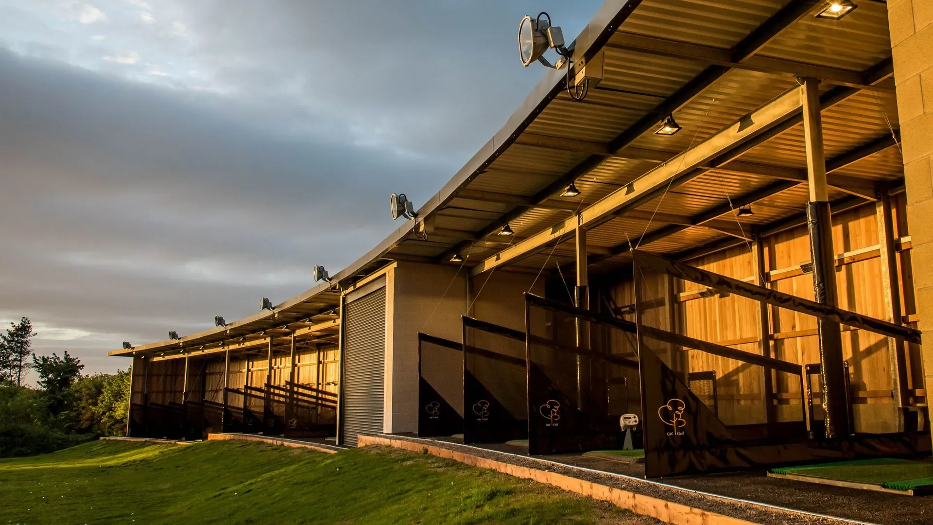 Covered golf driving range bays illuminated by warm evening light beneath a dramatic cloudy sky.