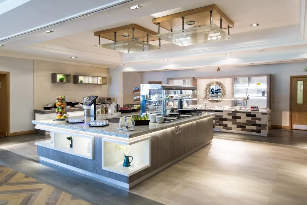 Modern hotel breakfast buffet area with granite counters, fruit display and illuminated shelving
