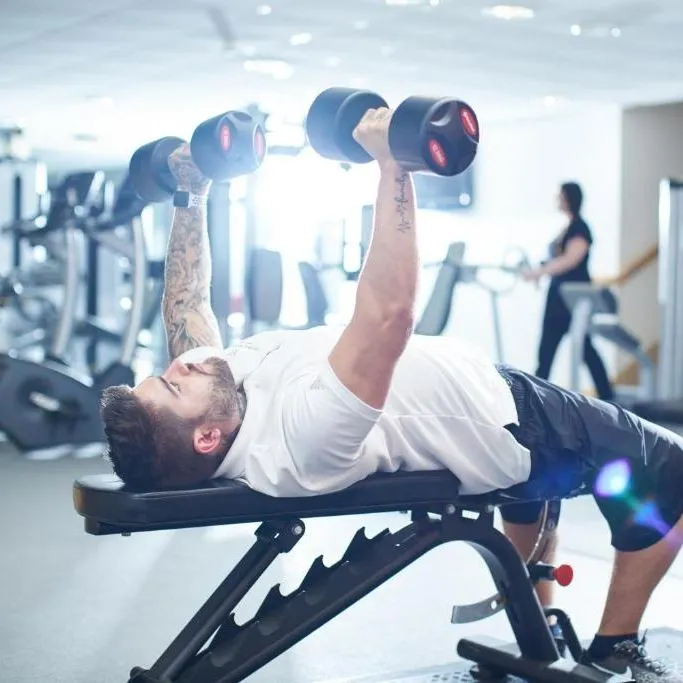 Tattooed man performing a dumbbell bench press on an adjustable bench in a bright gym.