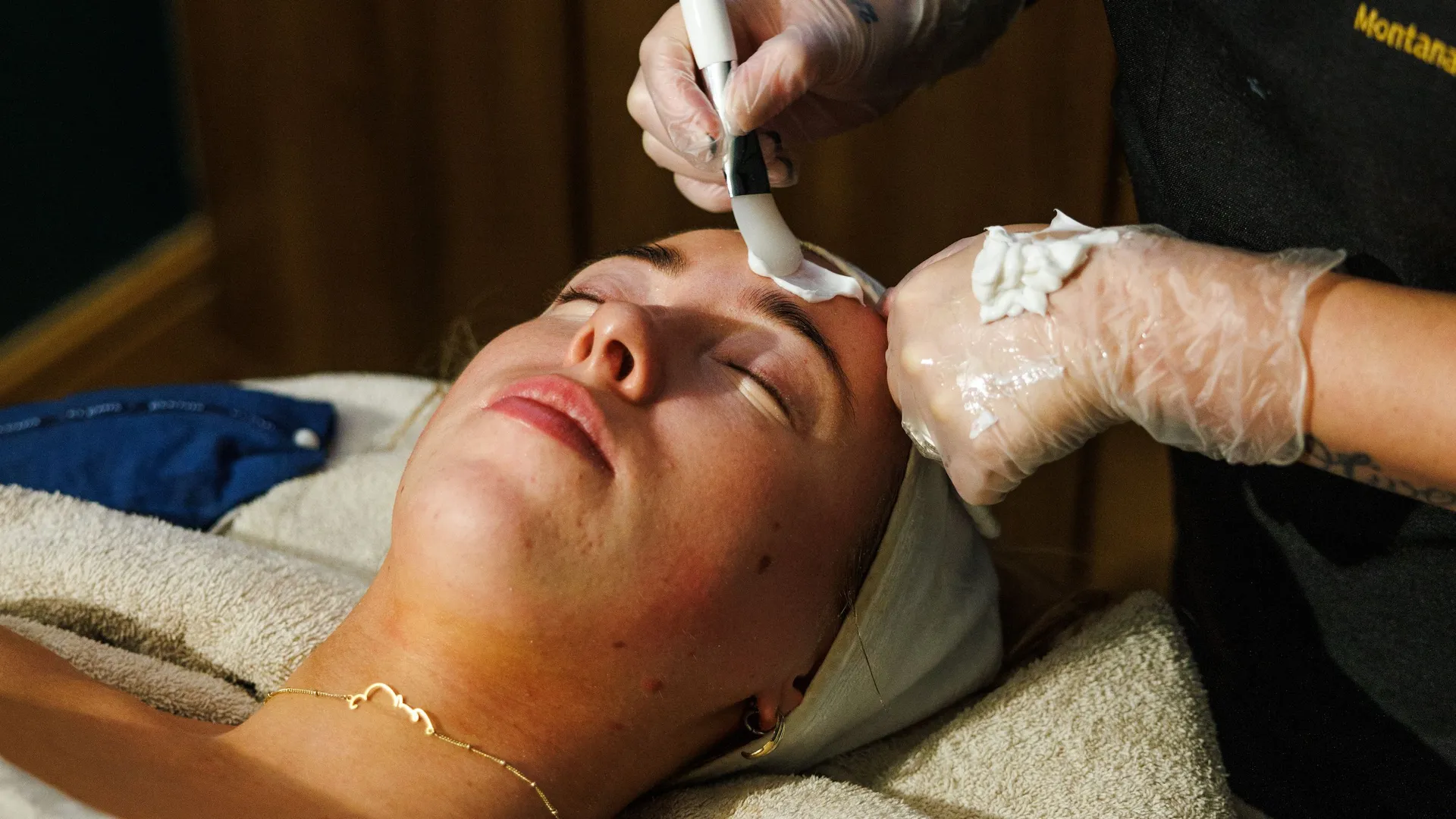 Beauty therapist applying a facial treatment with a brush to a reclining client's forehead.