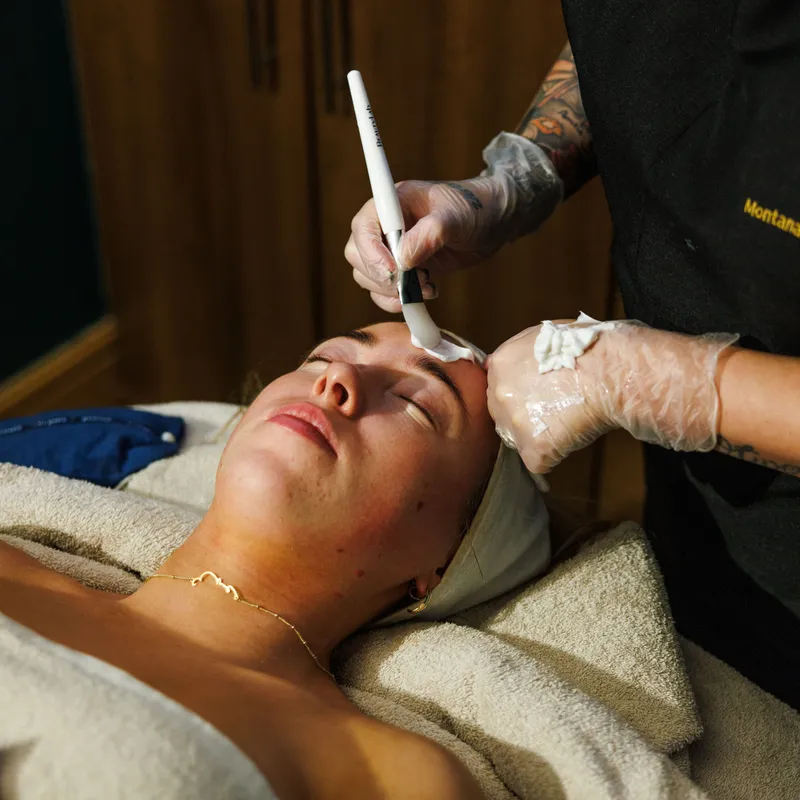 Beauty therapist applying a facial treatment with a brush to a reclining client's forehead.