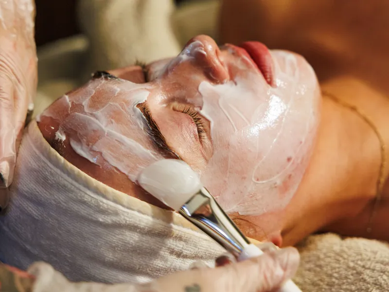 Person receiving a facial treatment with a brush applying white cream to their face.