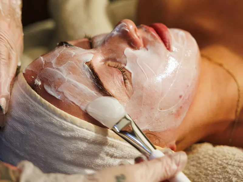 Person receiving a facial treatment with a brush applying white cream to their face.