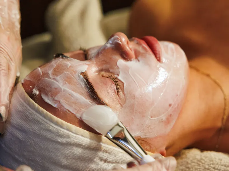 Person receiving a facial treatment with a brush applying white cream to their face.