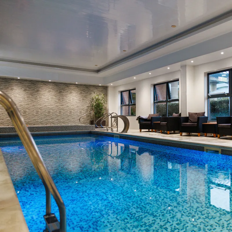 Modern indoor swimming pool with blue mosaic tiles, lounge chairs and large windows