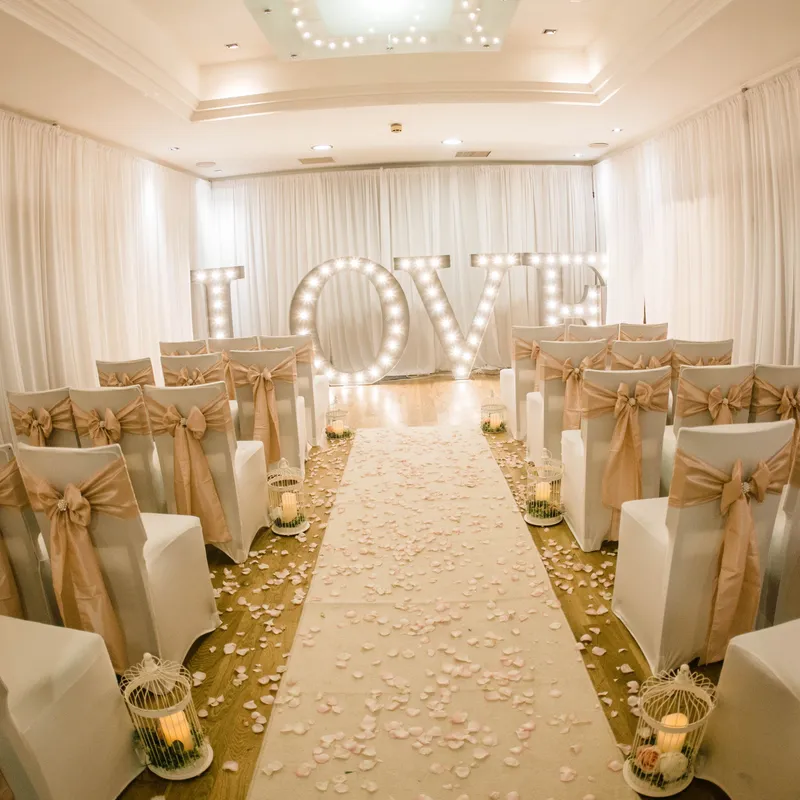 Elegant wedding ceremony room with white chair covers, gold bows, rose-petal aisle and illuminated LOVE letters.