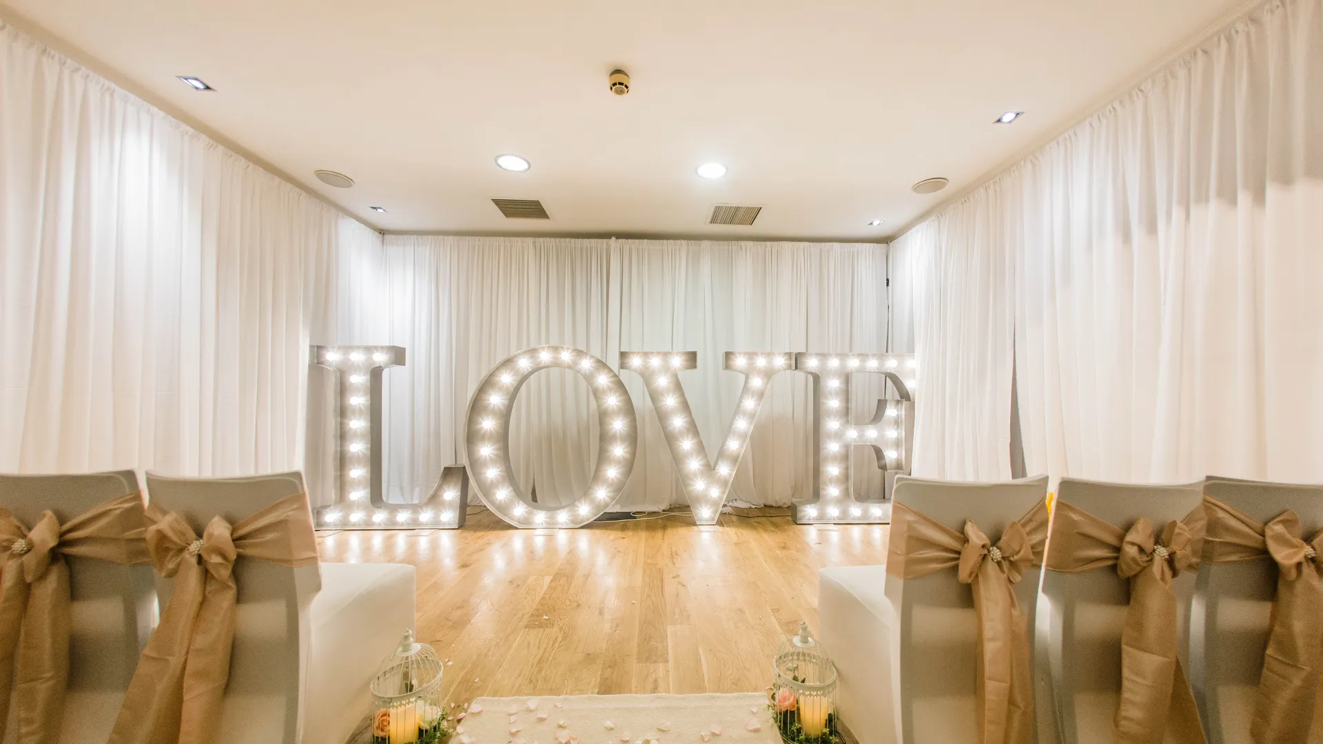 Elegant ceremony room with illuminated LOVE letters, white drapes and aisle chairs decorated with gold bows.
