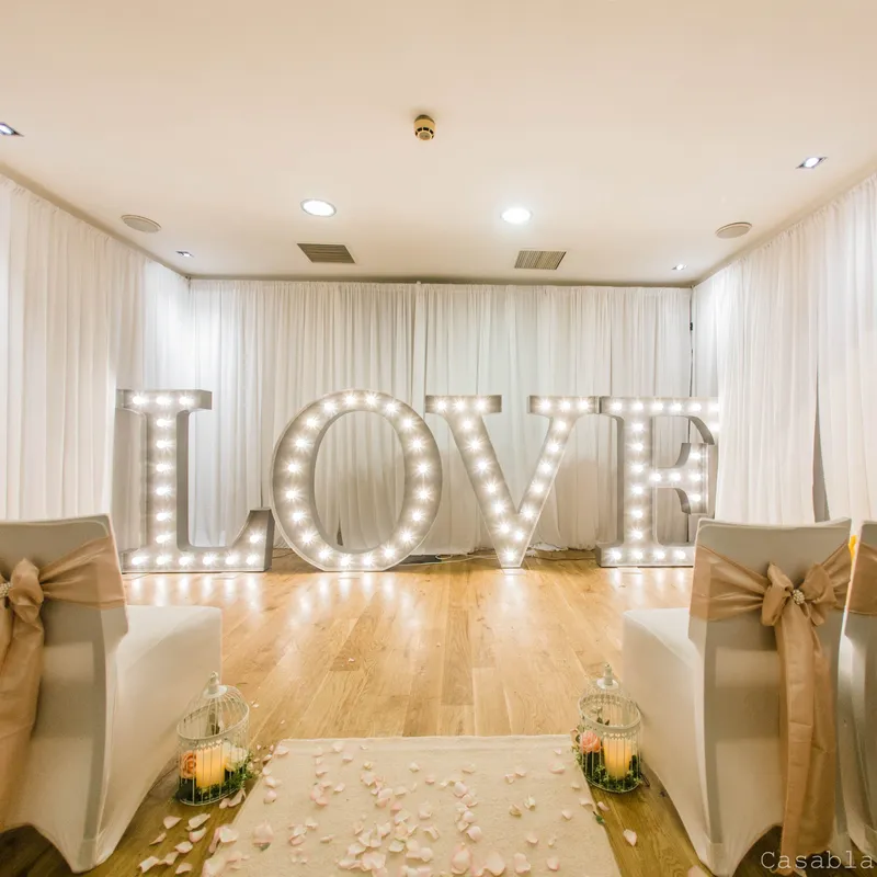 Elegant ceremony room with illuminated LOVE letters, white drapes and aisle chairs decorated with gold bows.