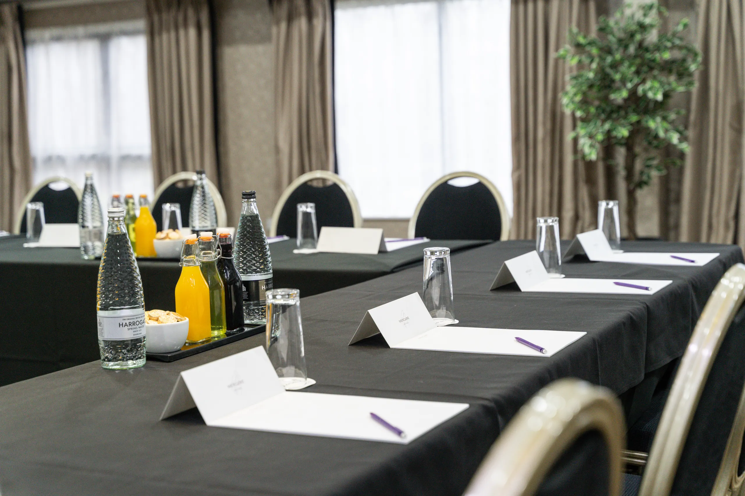 Conference room arranged with black-clothed tables, bottled water, juice, glasses, notepads and pens.