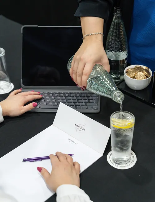 Person pouring sparkling water into a lemon drink beside a laptop and notebook