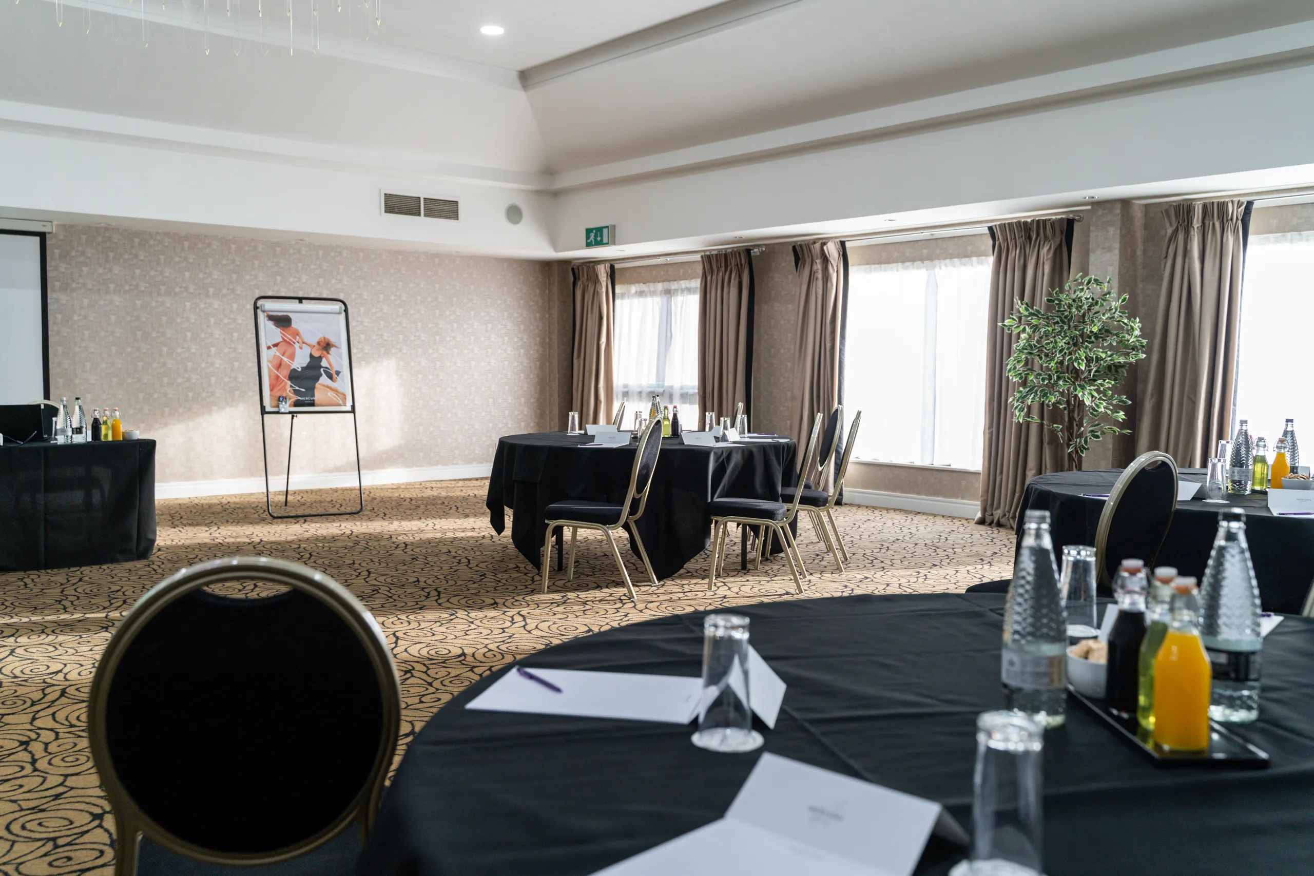 Bright hotel meeting room arranged with round black-clothed tables, chairs, bottled drinks and notepads.