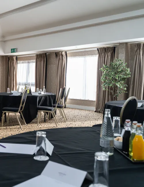 Bright hotel meeting room arranged with round black-clothed tables, chairs, bottled drinks and notepads.