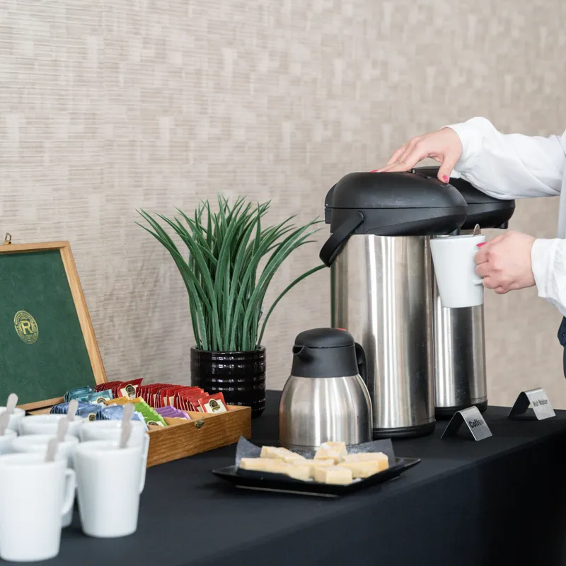 Woman pouring coffee from a stainless-steel airpot at a refreshment station with mugs, tea sachets and biscuits.