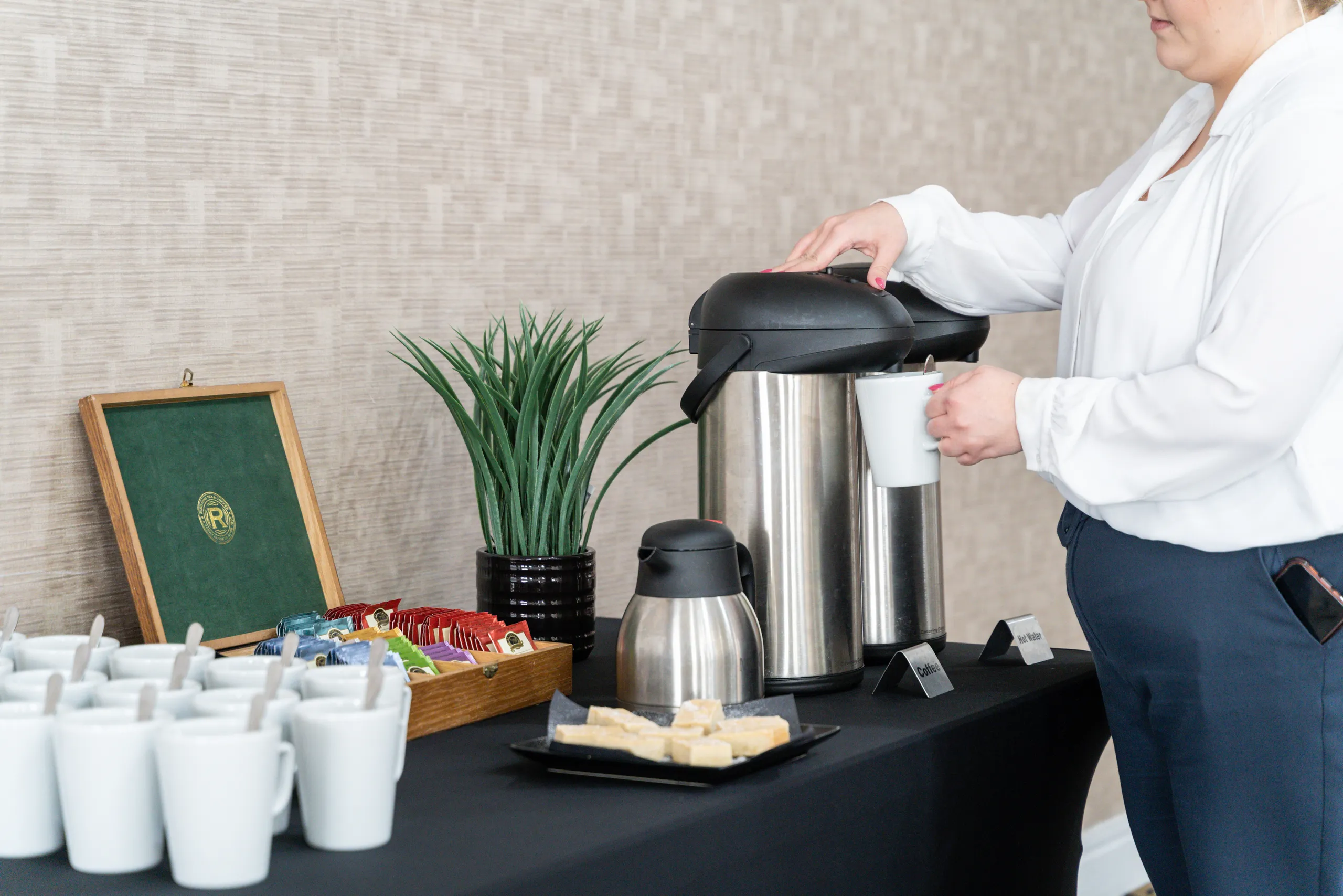 Woman pouring coffee from a stainless-steel airpot at a refreshment station with mugs, tea sachets and biscuits.