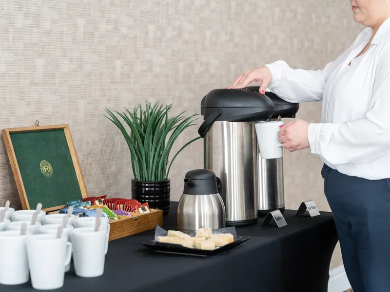 Woman pouring coffee from a stainless-steel airpot at a refreshment station with mugs, tea sachets and biscuits.