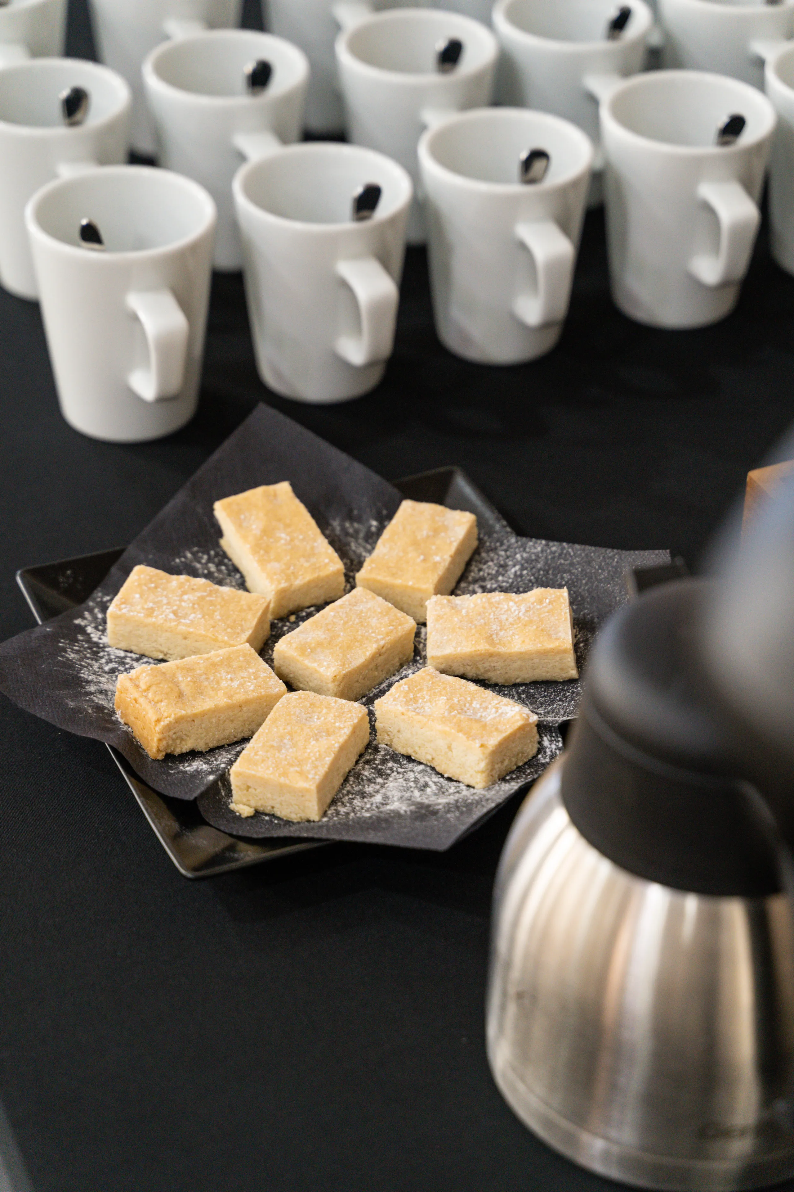 White mugs with teaspoons, shortbread biscuits and a stainless-steel coffee pot on a buffet table
