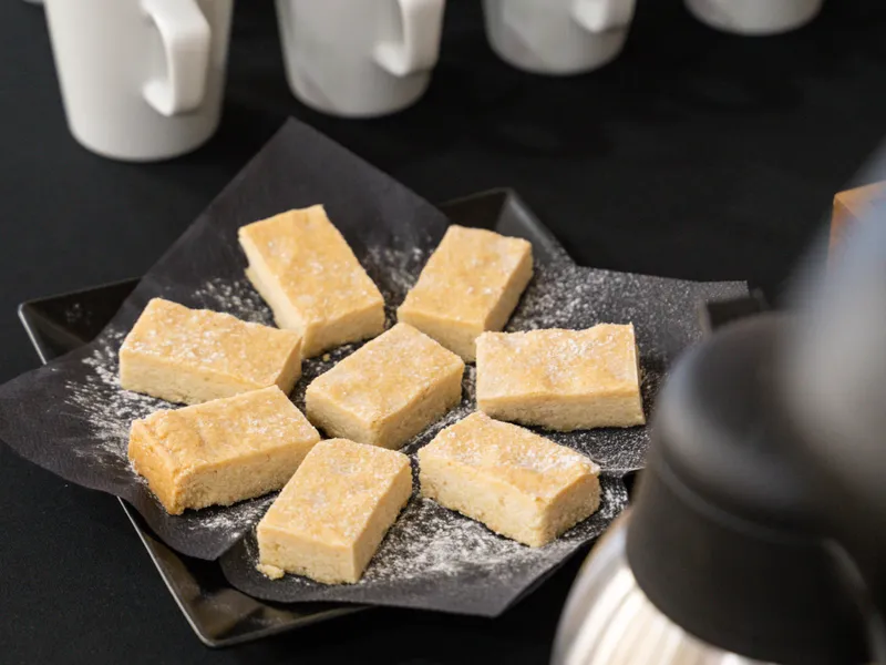 White mugs with teaspoons, shortbread biscuits and a stainless-steel coffee pot on a buffet table