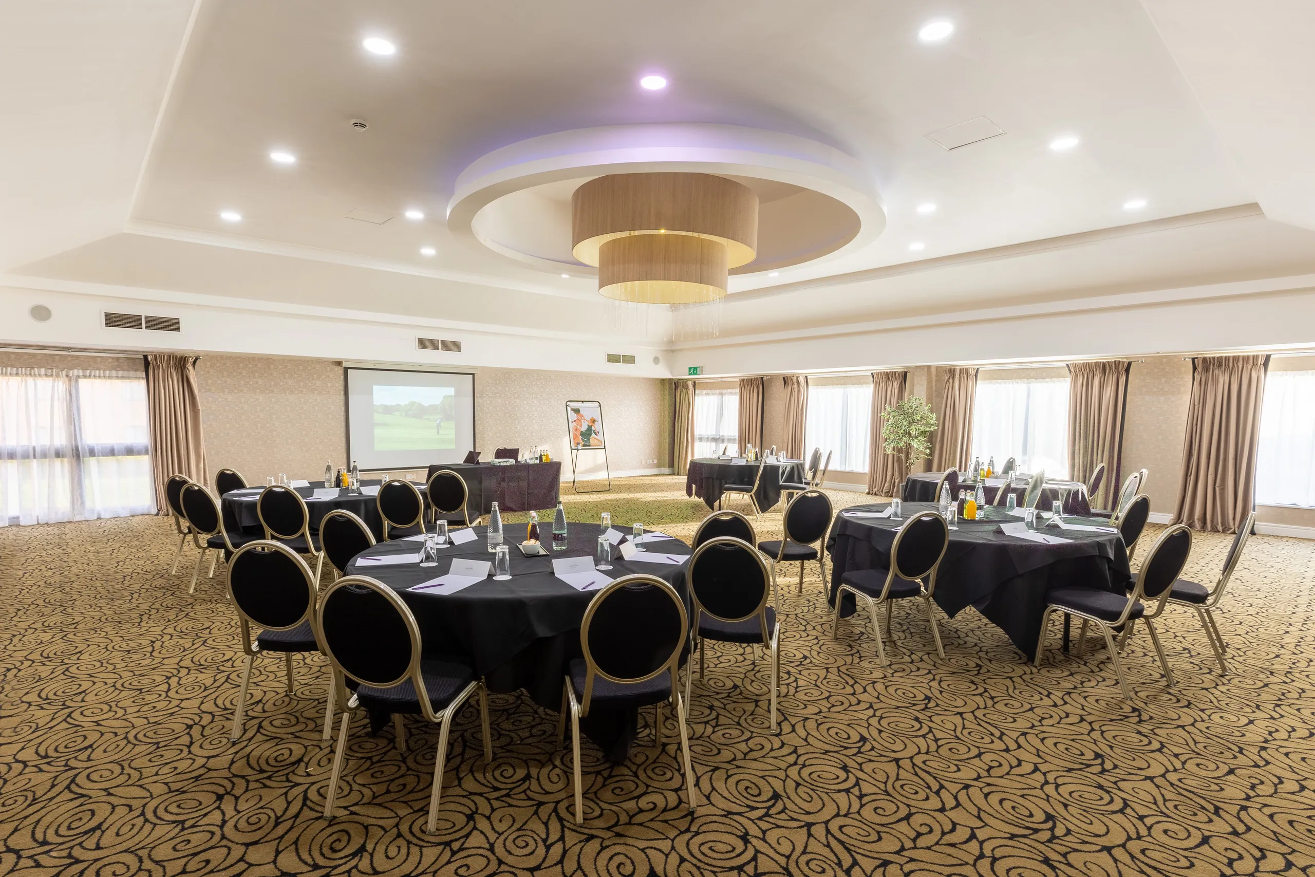 Elegant hotel conference room arranged with round tables, black chairs and a projector screen.