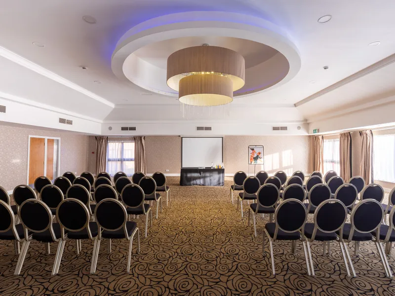 Conference room arranged with rows of black chairs facing a presentation screen beneath a modern circular chandelier.