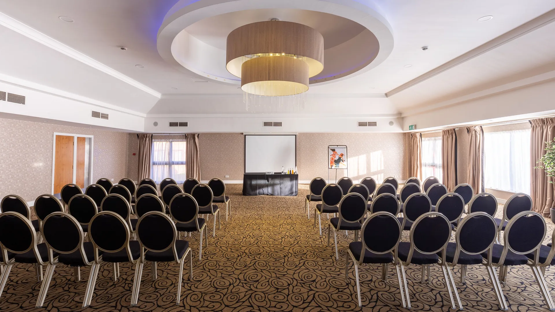 Conference room arranged with rows of black chairs facing a presentation screen beneath a modern circular chandelier.