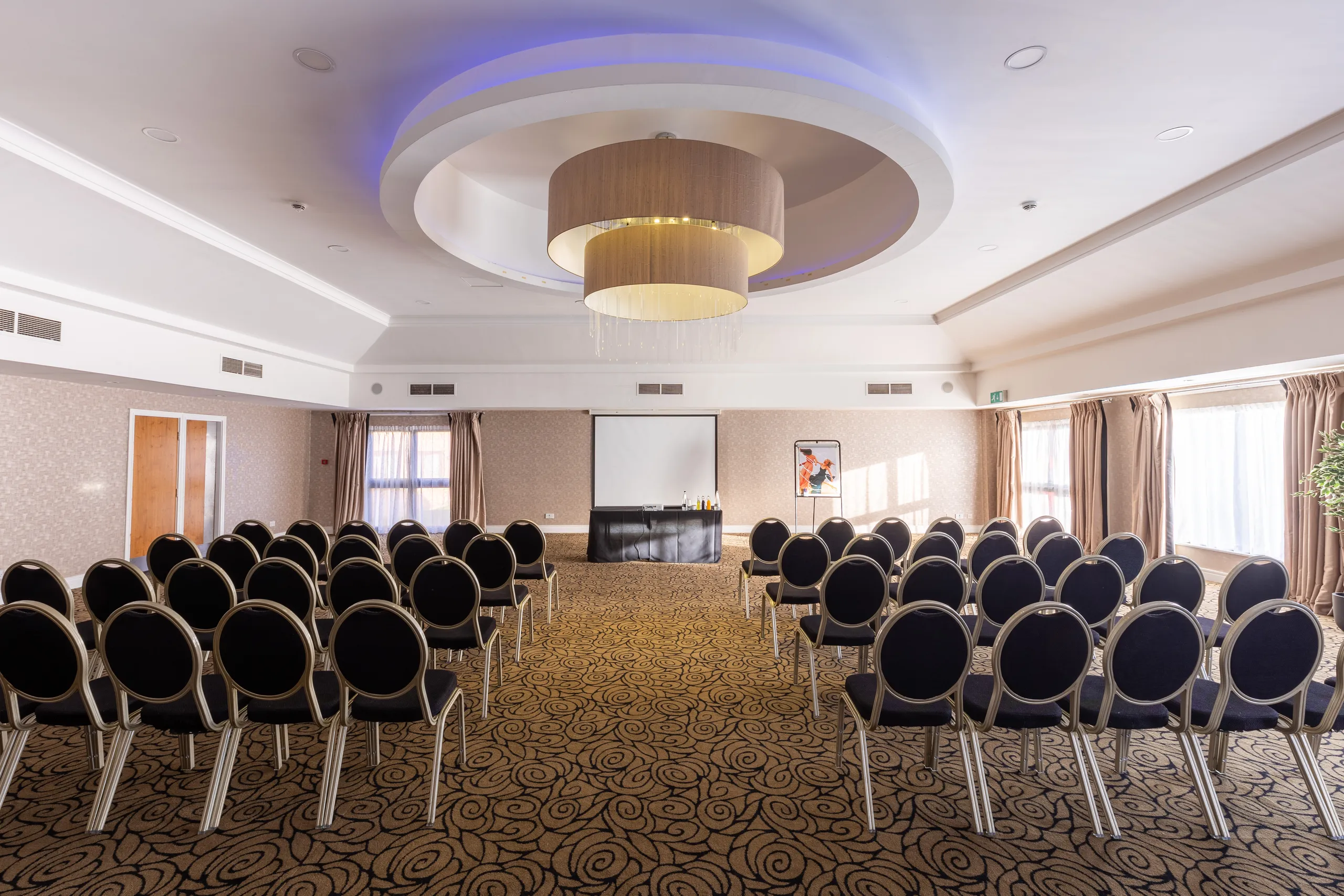 Conference room arranged with rows of black chairs facing a presentation screen beneath a modern circular chandelier.