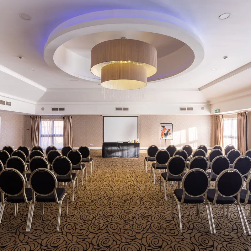 Conference room arranged with rows of black chairs facing a presentation screen beneath a modern circular chandelier.