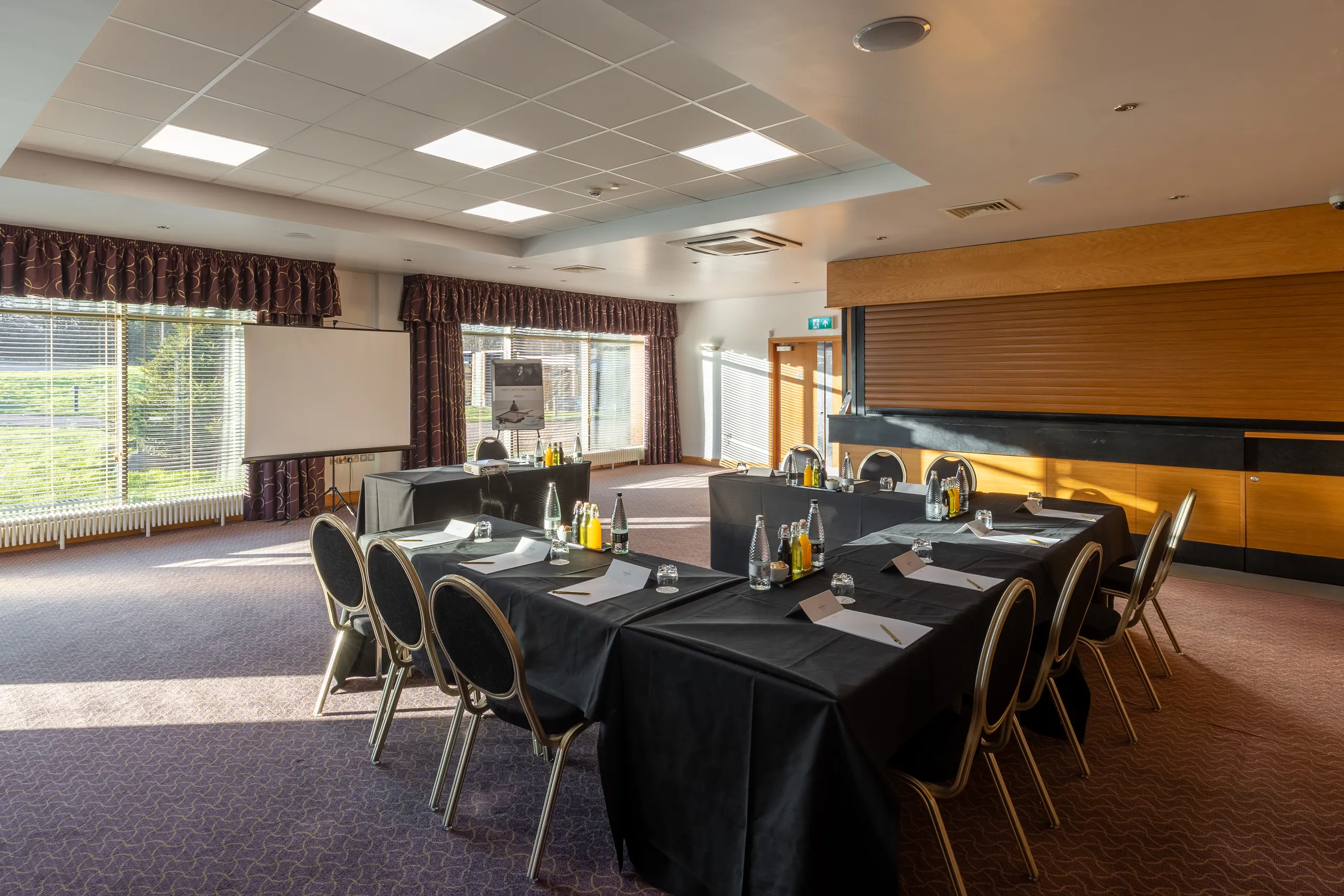 Hotel meeting room arranged in a U-shape with black-clothed tables, chairs, water bottles and presentation screen.