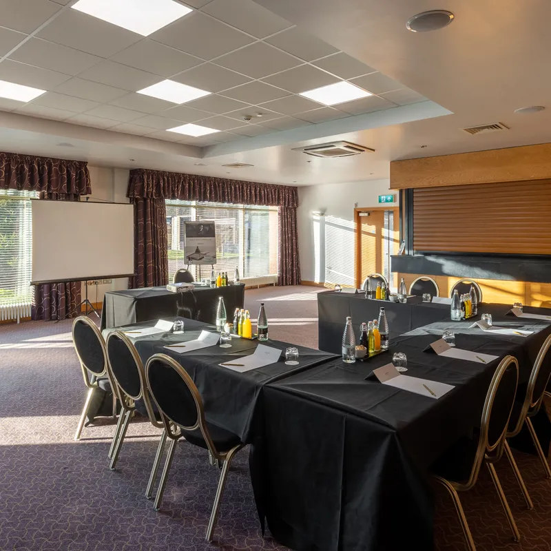 Hotel meeting room arranged in a U-shape with black-clothed tables, chairs, water bottles and presentation screen.