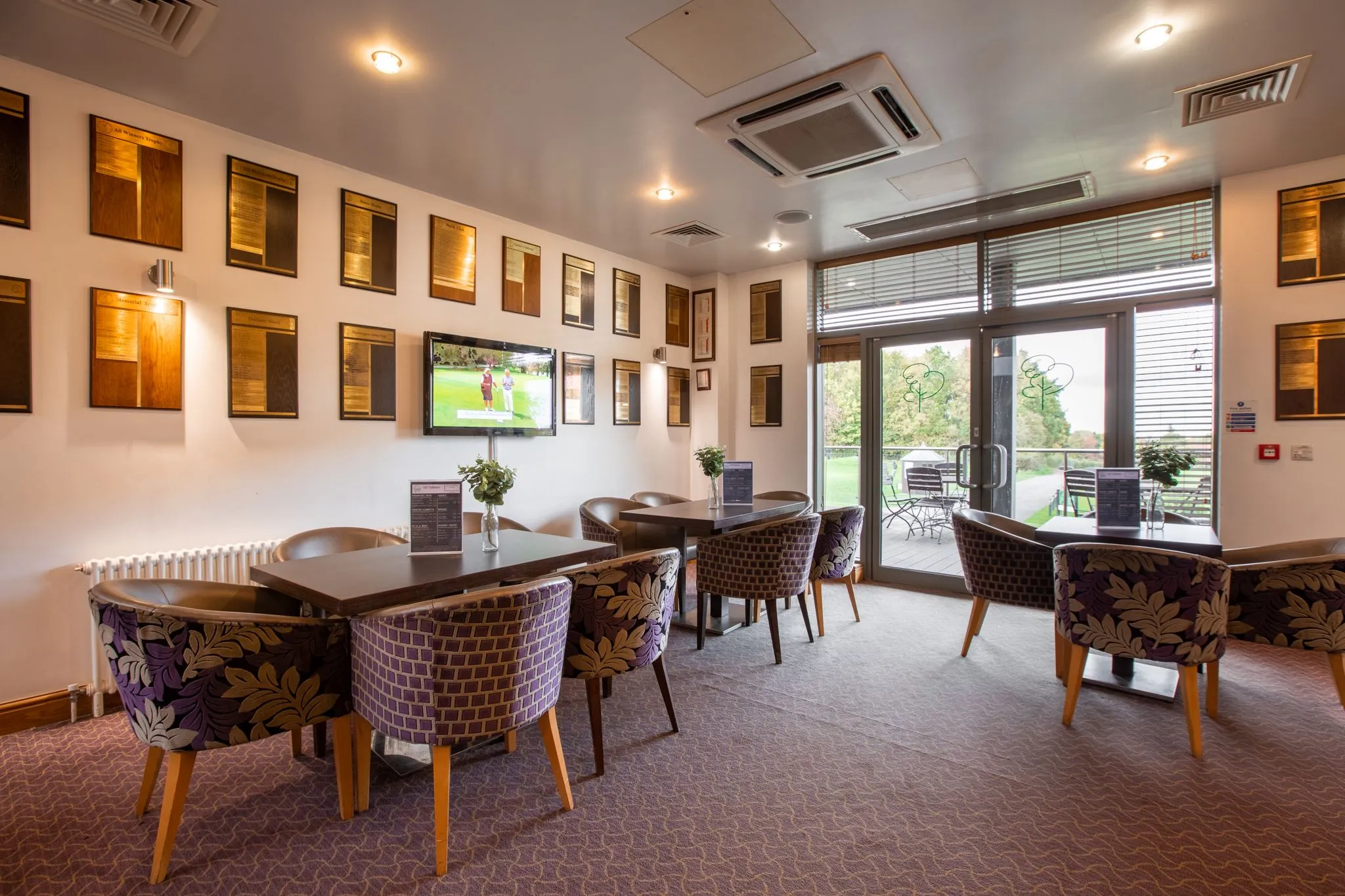 Elegant golf club lounge with patterned armchairs, dark wooden tables and views over a green through glass doors.
