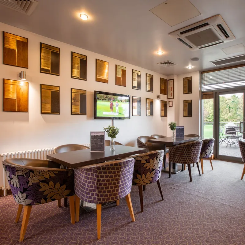 Elegant golf club lounge with patterned armchairs, dark wooden tables and views over a green through glass doors.