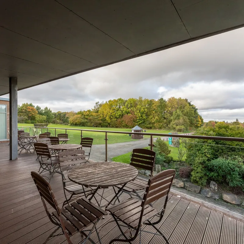 Covered terrace with wooden tables and folding chairs overlooking a green parkland landscape.