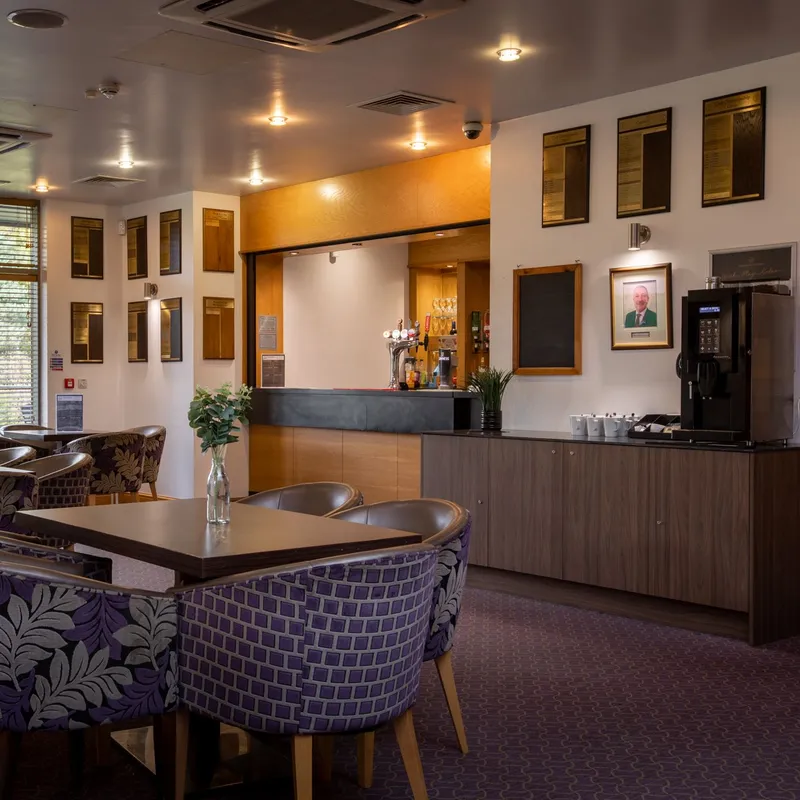 Warmly lit golf clubhouse lounge with patterned chairs, dining tables and a bar counter.